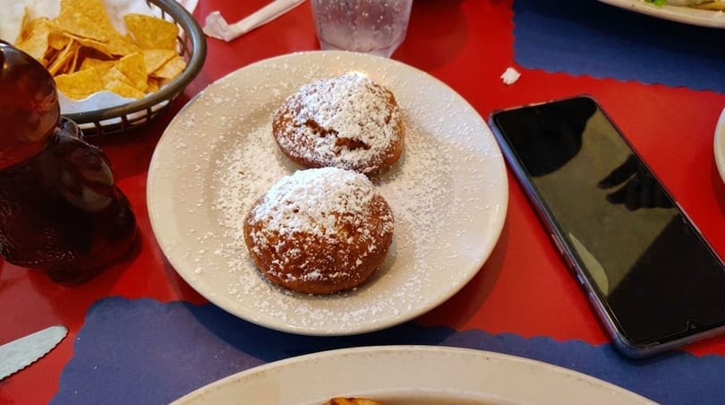 Two powdered sugar-dusted beignets on a white plate, with tortilla chips and a phone nearby.